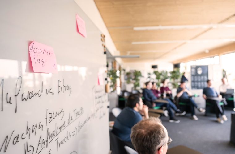 Meeting des AccsoNets in einem modernen, hellen Raum. Im Vordergrund steht ein Whiteboard mit Notizen, darunter "Accso Net-Tag," was auf ein technisches oder strategisches Thema hindeutet. Die Teilnehmer sitzen in einer informellen Runde, umgeben von Pflanzen.