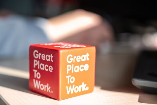 Ein roter und orangefarbener Würfel mit der Aufschrift ‚Great Place To Work‘ steht auf einem Schreibtisch. Die Auszeichnung symbolisiert ein ausgezeichnetes Arbeitsumfeld.