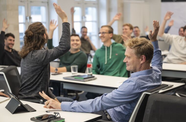 Eine Gruppe von Personen sitzt in einem Seminarraum an Tischen und hebt die Hände, möglicherweise um an einer Diskussion oder Abstimmung teilzunehmen. Der Raum ist hell erleuchtet mit großen Fenstern im Hintergrund. Auf den Tischen liegen Tablets und Notizblöcke, was auf eine moderne Lernumgebung hinweist.