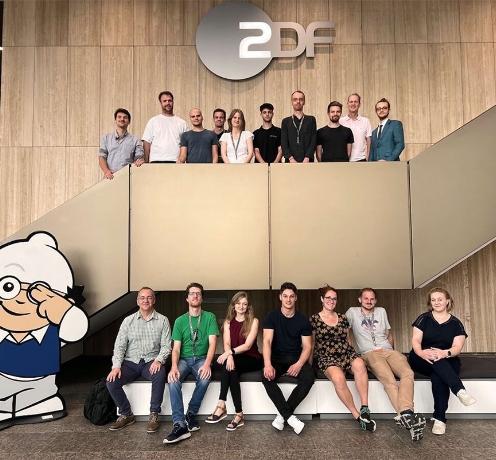 Das Bild zeigt eine Gruppe von Personen, die sich in einem modernen Gebäude versammelt haben. Sie stehen auf und um eine Treppe herum, mit einem großen "ZDF"-Logo an der Wand über ihnen. Im Vordergrund ist eine große Cartoon-Figur mit Brille und weißer Mütze zu sehen, die einen blauen Pullover und graue Hosen trägt und freundlich wink.