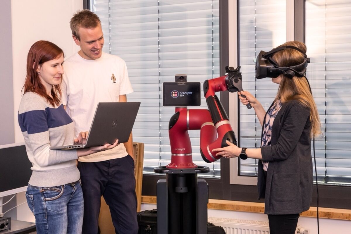 Drei Personen arbeiten in einem Büro mit einem roten Roboterarm namens "Sawyer". Eine Person trägt ein Virtual-Reality-Headset, eine andere hält einen Laptop. Die Umgebung ist technisch ausgestattet.