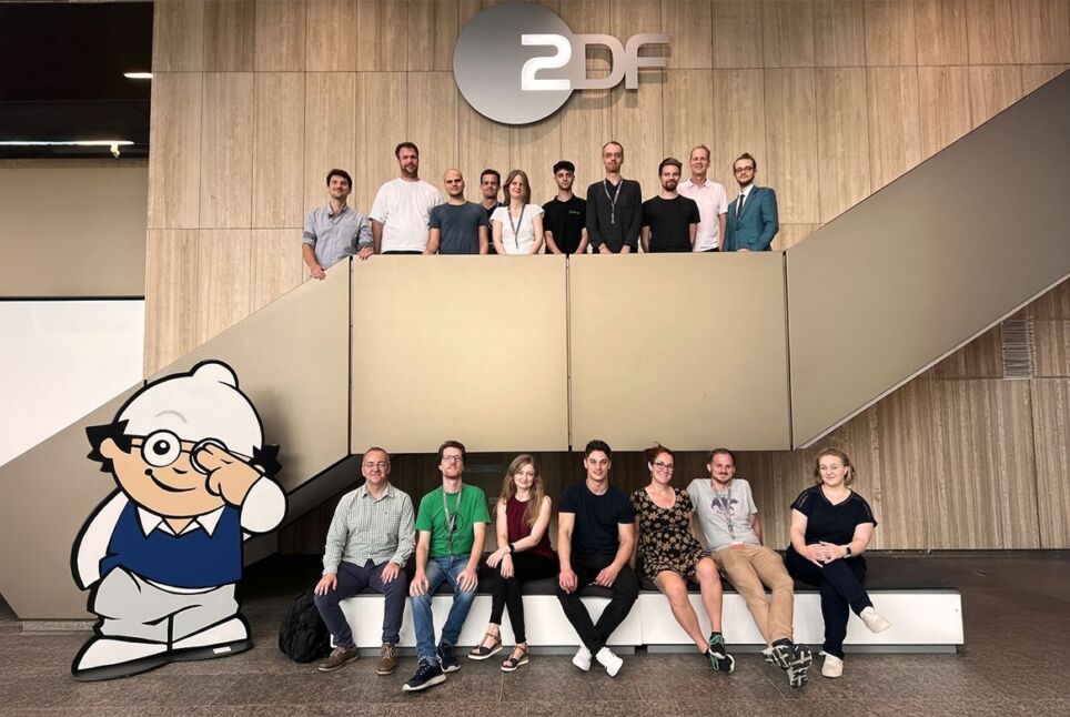 Das Bild zeigt eine Gruppe von Personen, die sich in einem modernen Gebäude versammelt haben. Sie stehen auf und um eine Treppe herum, mit einem großen "ZDF"-Logo an der Wand über ihnen. Im Vordergrund ist eine große Cartoon-Figur mit Brille und weißer Mütze zu sehen, die einen blauen Pullover und graue Hosen trägt und freundlich wink.