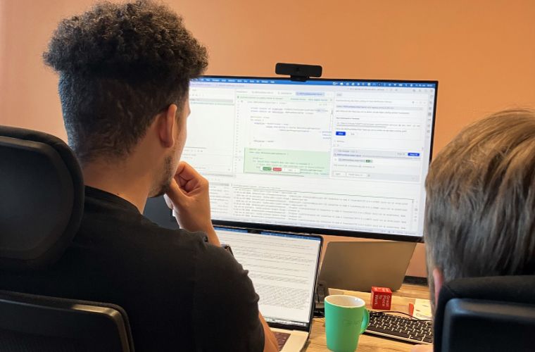 Zwei Personen sitzen von hinten an einem Schreibtisch; ein großer Monitor zeigt Code- und Softwarefenster, daneben Laptop, grüne Tasse und Notizen.