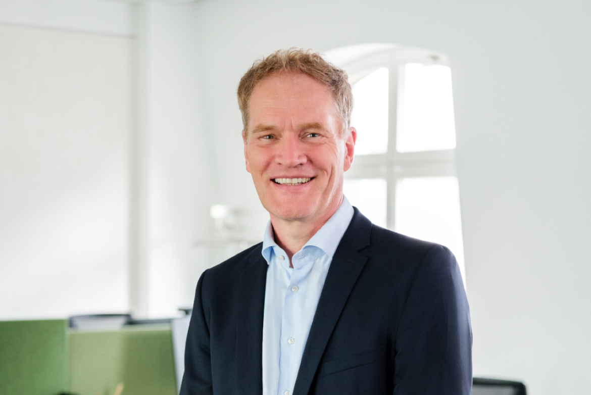 Person in dunklem Blazer und hellblauem Hemd lächelt in einem hellen Büro; Hintergrund mit großem Fenster und grünen Trennwänden.