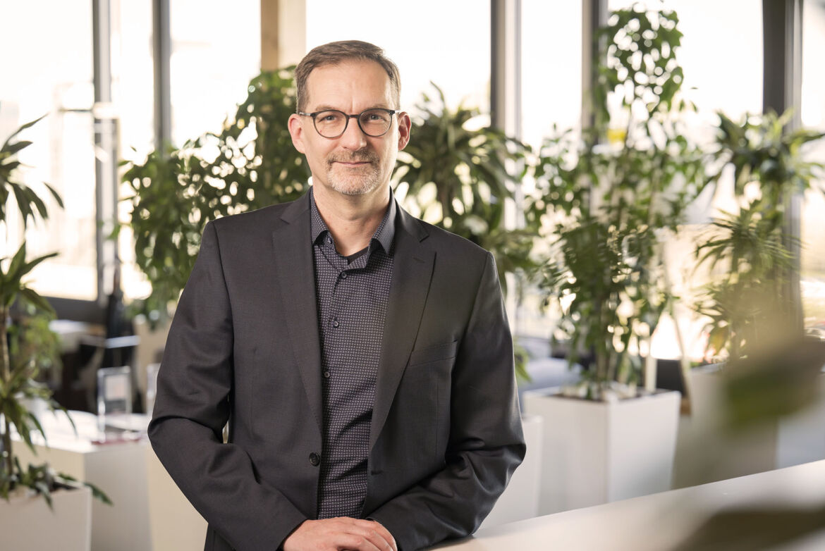 Thomas Jäger in einem dunklen Anzug, die in einer modernen Büroumgebung steht. Im Hintergrund sind große Fenster und mehrere grüne Pflanzen zu sehen, was eine helle und natürliche Atmosphäre schafft.