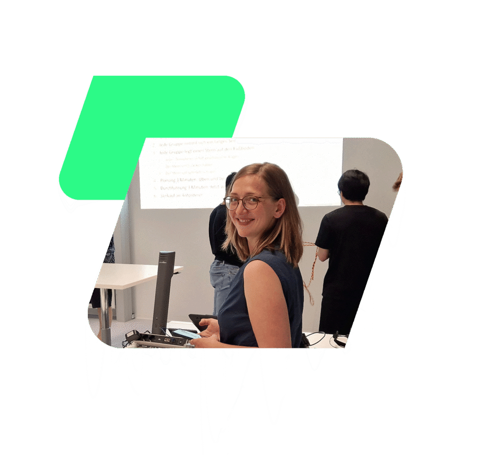 Eine Person steht in einem Raum vor einem Projektorbildschirm, auf dem Text angezeigt wird. Im Hintergrund sind weitere Personen zu sehen, die sich dem Bildschirm zuwenden. Im Vordergrund befindet sich ein Tisch mit technischen Geräten.