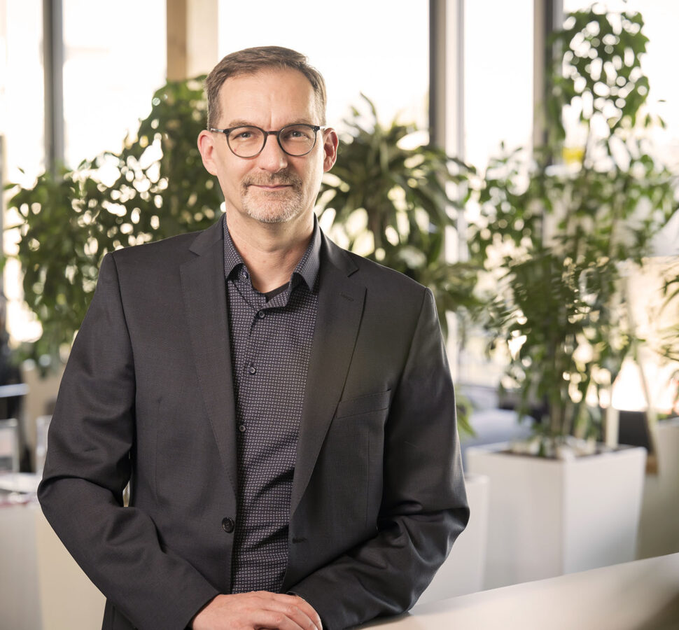 Thomas Jäger in einem dunklen Anzug, die in einer modernen Büroumgebung steht. Im Hintergrund sind große Fenster und mehrere grüne Pflanzen zu sehen, was eine helle und natürliche Atmosphäre schafft.