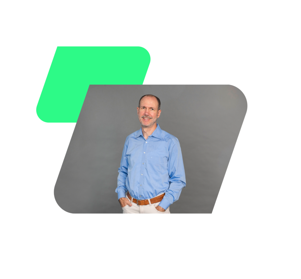 Eine Person in einem blauen Hemd und beigen Hosen steht mit den Händen in den Taschen vor einem grauen Hintergrund, eingerahmt von einem stilisierten grünen und schwarzen geometrischen Design.