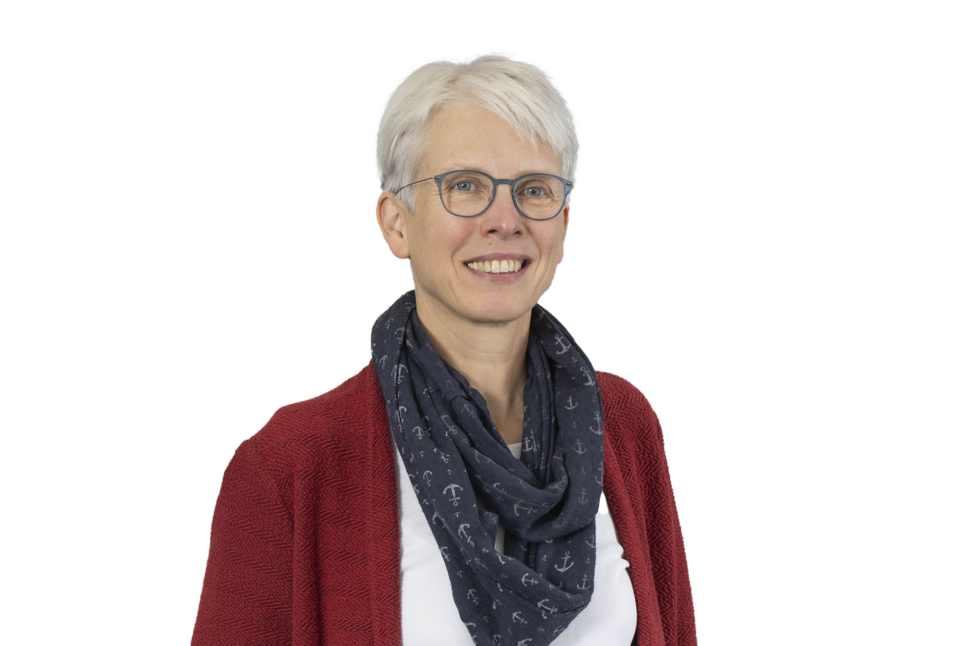 Porträt einer Frau mit kurzen weißen Haaren, Brille und einem dunklen Schal mit Anker-Muster. Sie trägt eine rote Strickjacke über einem weißen Oberteil und lächelt. Der Hintergrund ist grau.