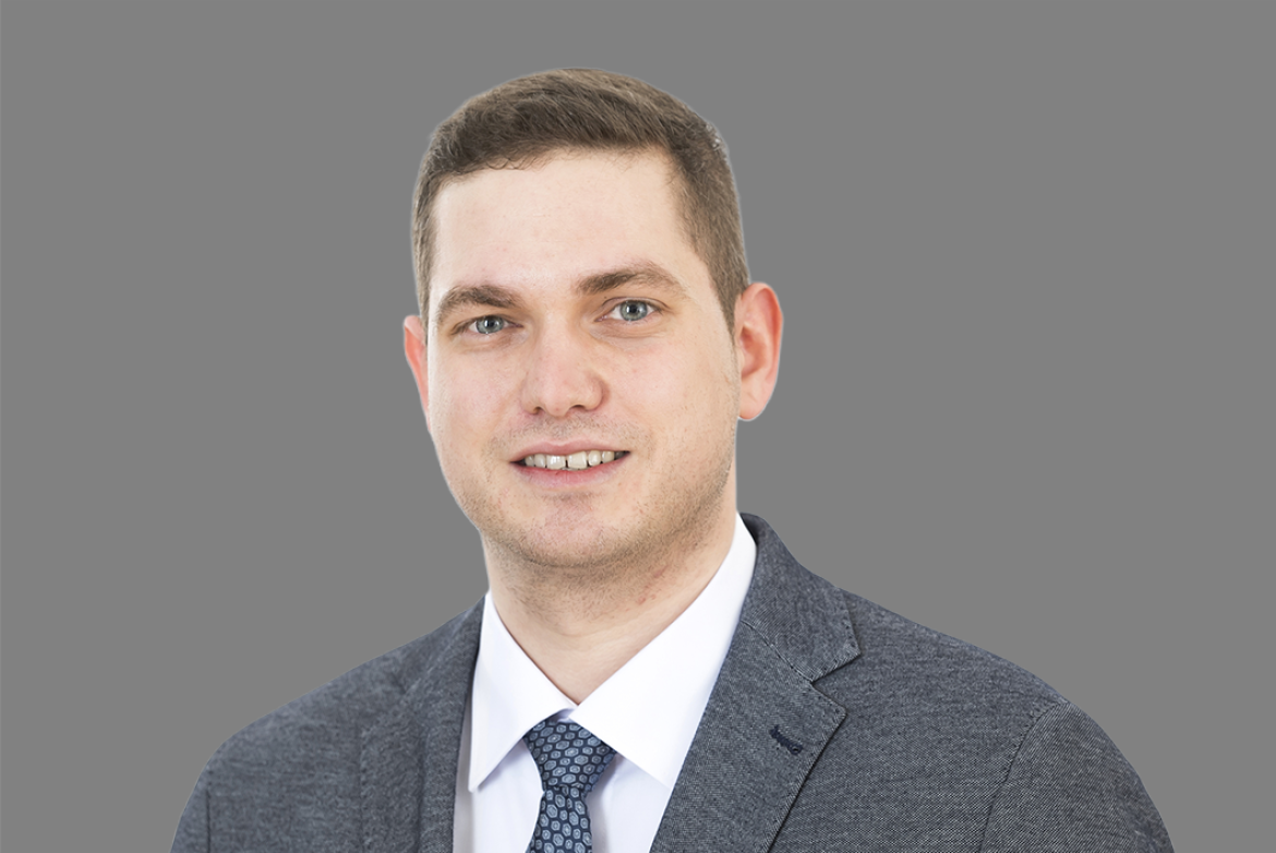 Porträt einer Person in grauem Anzug, weißem Hemd und einer gemusterten Krawatte vor grauem Hintergrund.