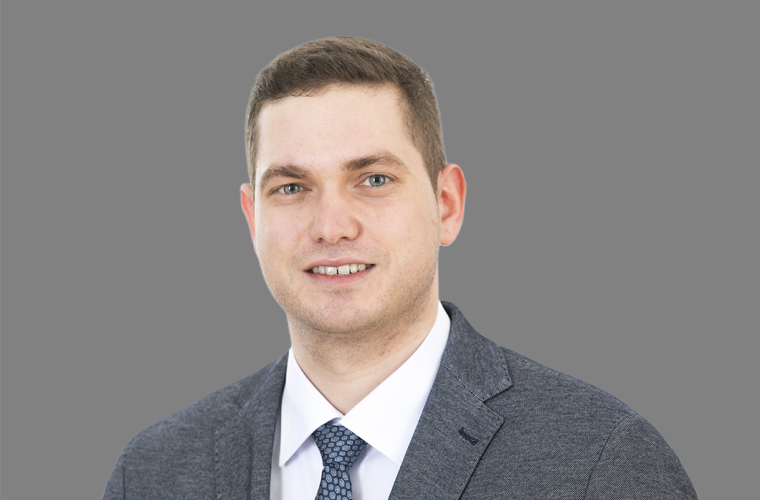 Porträt einer Person in grauem Anzug, weißem Hemd und einer gemusterten Krawatte vor grauem Hintergrund.