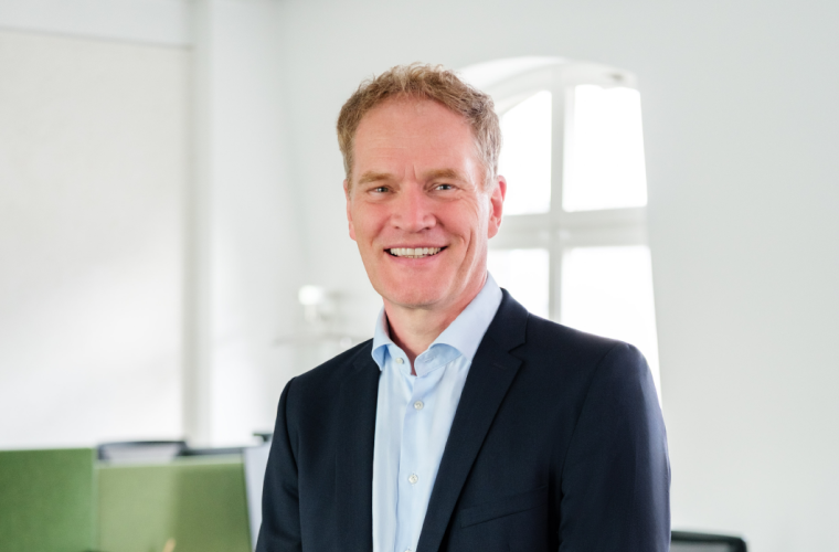 Person in dunklem Blazer und hellblauem Hemd lächelt in einem hellen Büro; Hintergrund mit großem Fenster und grünen Trennwänden.