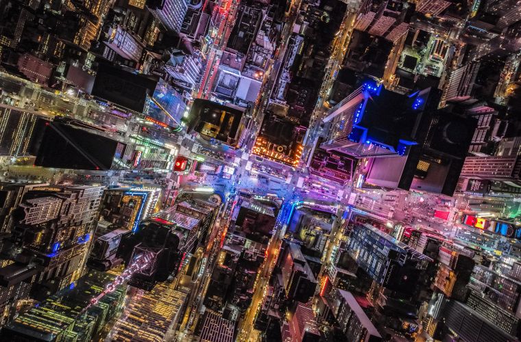 Eine nächtliche Luftaufnahme einer Stadt, die von einem Netz aus Straßen und beleuchteten Gebäuden durchzogen ist. Die Lichter der Stadt bilden ein komplexes Muster aus gelben und weißen Linien, die die Straßen und Gebäudeumrisse nachzeichnen. Dunkle Bereiche zwischen den beleuchteten Zonen zeigen unbebaute oder weniger beleuchtete Gebiete.