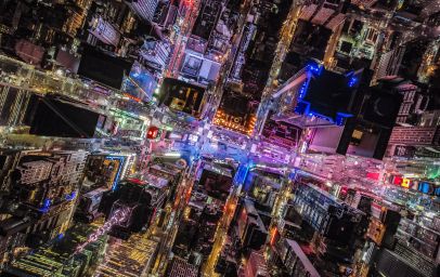 Eine nächtliche Luftaufnahme einer Stadt, die von einem Netz aus Straßen und beleuchteten Gebäuden durchzogen ist. Die Lichter der Stadt bilden ein komplexes Muster aus gelben und weißen Linien, die die Straßen und Gebäudeumrisse nachzeichnen. Dunkle Bereiche zwischen den beleuchteten Zonen zeigen unbebaute oder weniger beleuchtete Gebiete.
