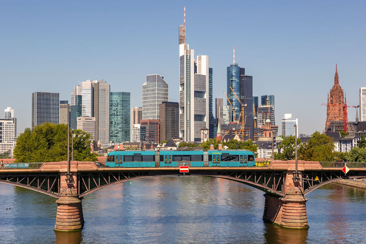 Ein moderner Zug fährt über eine Brücke in einer Stadt mit einer beeindruckenden Skyline. Im Hintergrund sind die Wolkenkratzer von Frankfurt am Main zu sehen, darunter der Commerzbank Tower und andere hohe Gebäude. Der Fluss im Vordergrund spiegelt die Architektur wider, und der Himmel ist klar und blau. Auf der rechten Seite ist der historische Kaiserdom St. Bartholomäus zu erkennen.