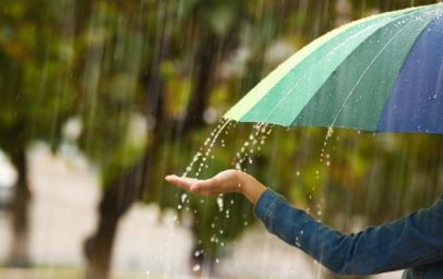 Eine Person, die einen mehrfarbigen Regenschirm hält, während es regnet. Eine Hand wird unter den Schirm ausgestreckt, um die fallenden Regentropfen zu spüren. Im Hintergrund sind verschwommene grüne Bäume zu sehen.