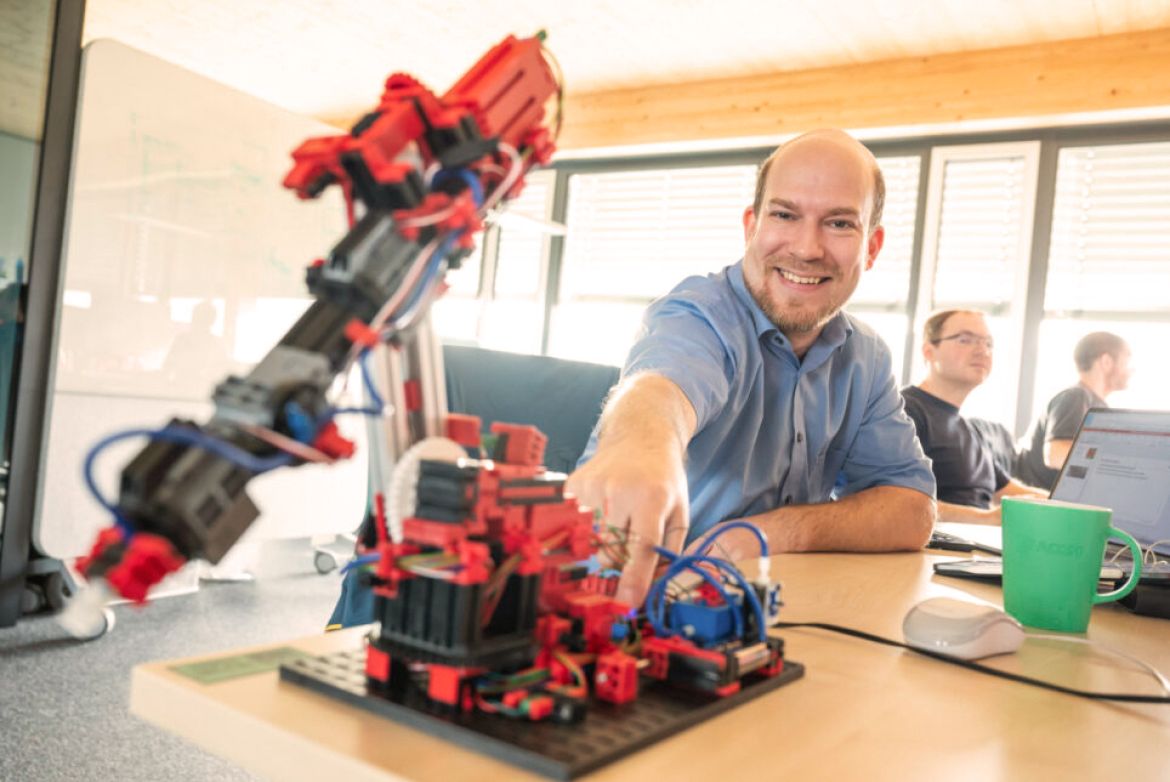 Ein Mann im blauen Hemd lacht in die Kamera. Vor ihm steht ein mit Fischer Technik gebauter Roboter, bei welchem er auf einen Knopf drückt.