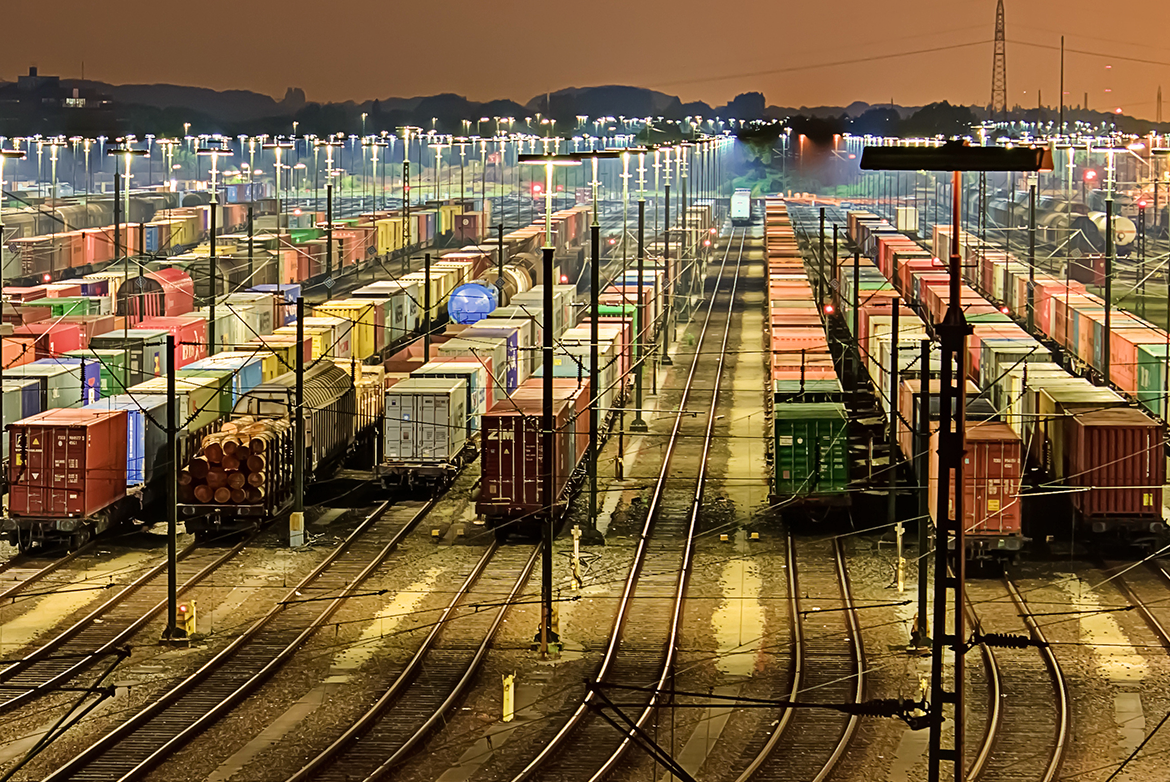 Das Bild zeigt einen nächtlichen Güterbahnhof mit vielen beleuchteten Gleisen. Zahlreiche bunte Frachtcontainer in Rot, Grün, Blau und Gelb stehen auf den Gleisen. Im Hintergrund sind Bäume und Gebäude silhouettenhaft vor einem orangefarbenen Himmel zu sehen.