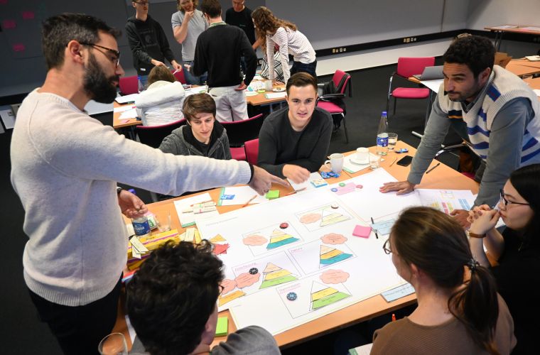 Eine Gruppe von Personen arbeitet in einem Workshop an einem Tisch mit großen Plakaten, Diagrammen und Notizzetteln. Eine Person zeigt auf eine Stelle auf dem Plakat, während die anderen aufmerksam zuhören und diskutieren. Im Hintergrund arbeiten weitere Gruppen an ähnlichen Aufgaben.