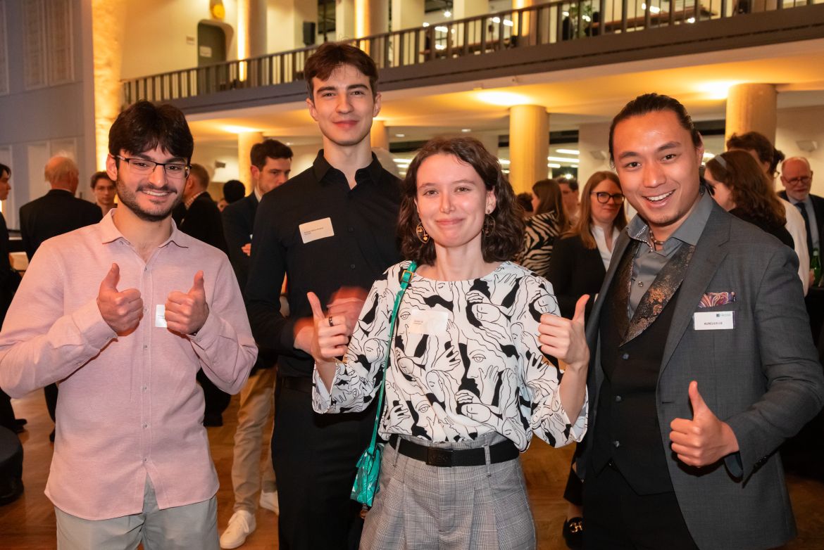 Die drei von Accso geförderten Deutschlandstipendiat:innen stehen mit einem Accso-Vertreter auf der Stipendienfeier zusammen und zeigen Daumen hoch; im Hintergrund weitere Gäste.