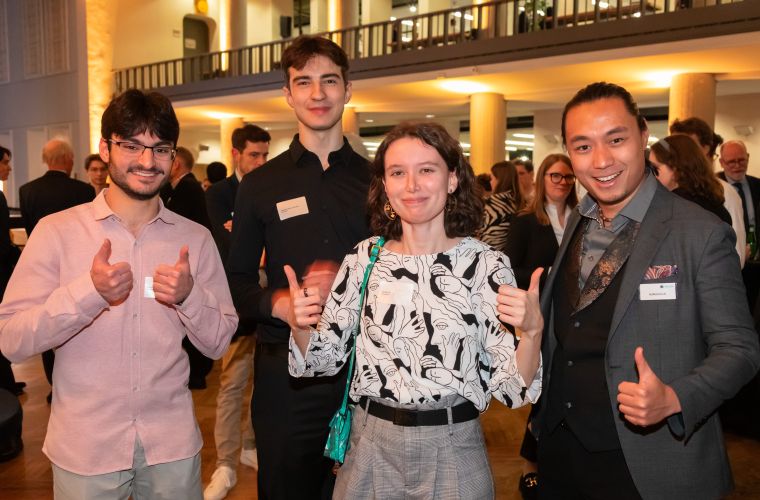 Die drei von Accso geförderten Deutschlandstipendiat:innen stehen mit einem Accso-Vertreter auf der Stipendienfeier zusammen und zeigen Daumen hoch; im Hintergrund weitere Gäste.